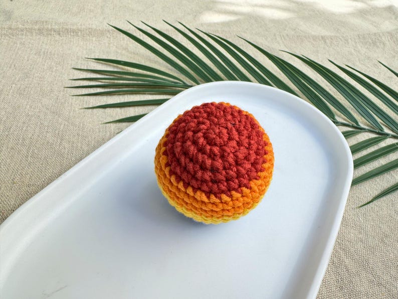 K&ouml;nnte beinhalten: Ein handgeh&auml;kelter Ball in Orange- und Gelbt&ouml;nen, der auf einem wei&szlig;en, ovalen Teller liegt. Die Textur des Balls ist sichtbar, mit einem Farbverlauf. Ein gr&uuml;nes Palmenblatt befindet sich im Hintergrund.