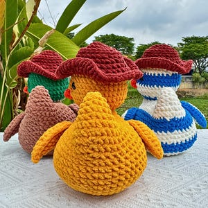 Pu&ograve; includere: Un gruppo di anatroccoli all'uncinetto fatti a mano in vari colori. Gli anatroccoli hanno cappelli rossi e sono nelle tonalit&agrave; del giallo, marrone, verde e blu. Gli anatroccoli sono su una superficie bianca con fogliame verde sullo sfondo.