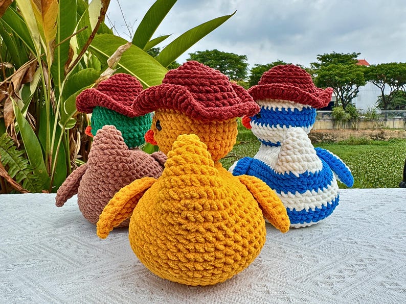 Pu&ograve; includere: Un gruppo di anatroccoli all'uncinetto fatti a mano in vari colori. Gli anatroccoli hanno cappelli rossi e sono nelle tonalit&agrave; del giallo, marrone, verde e blu. Gli anatroccoli sono su una superficie bianca con fogliame verde sullo sfondo.