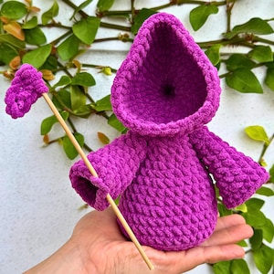 May include: A purple crocheted hooded doll with a stick in its hand. The doll is sitting in a person's hand.