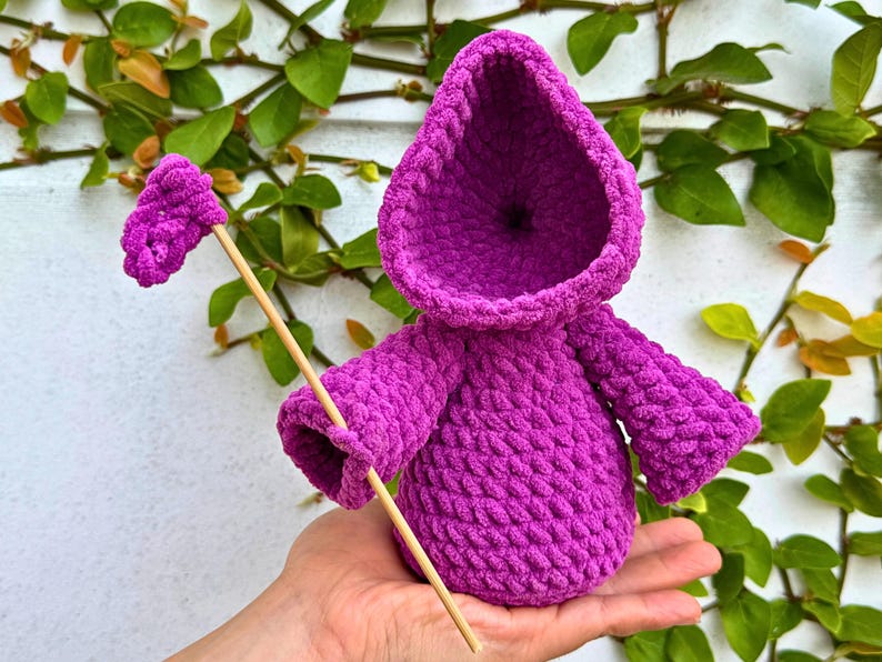 May include: A purple crocheted hooded doll with a stick in its hand. The doll is sitting in a person's hand.