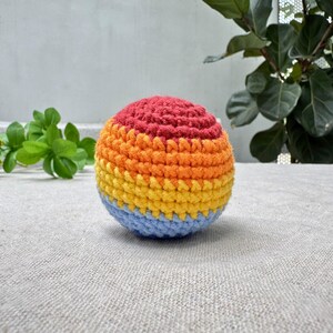 K&ouml;nnte beinhalten: Ein mehrfarbiger geh&auml;kelter Ball mit B&auml;ndern in Rot, Orange, Gelb und Blau. Der handgefertigte Ball hat eine strukturierte Oberfl&auml;che. Der Ball liegt auf einer hellgrauen Oberfl&auml;che, mit gr&uuml;nen Pflanzen im Hintergrund.