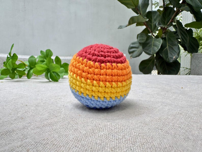 K&ouml;nnte beinhalten: Ein mehrfarbiger geh&auml;kelter Ball mit B&auml;ndern in Rot, Orange, Gelb und Blau. Der handgefertigte Ball hat eine strukturierte Oberfl&auml;che. Der Ball liegt auf einer hellgrauen Oberfl&auml;che, mit gr&uuml;nen Pflanzen im Hintergrund.