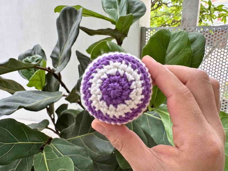 K&ouml;nnte beinhalten: Ein geh&auml;kelter Ball mit einem Spiralmuster in Lila und Wei&szlig;. Die handgefertigte Kugel wird vor einer gr&uuml;nen, belaubten Pflanze gehalten. Der Ball hat einen Durchmesser von etwa 6,35 cm.