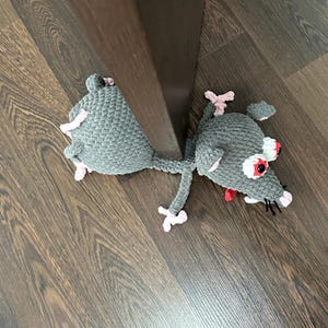 Puede incluir: Un tope de puerta de ratón gris tejido a mano con detalles en rosa. El ratón tiene grandes ojos rojos y blancos, orejas rosas y una nariz rosa. El tope de puerta está sobre un suelo de madera.