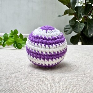 K&ouml;nnte beinhalten: Handgeh&auml;kelter Ball in Kugelform mit abwechselnden wei&szlig;en und lila Streifen. Der Ball liegt auf einer neutralfarbenen Oberfl&auml;che, im Hintergrund gr&uuml;nes Laub. Der Durchmesser betr&auml;gt ca. 7 cm.