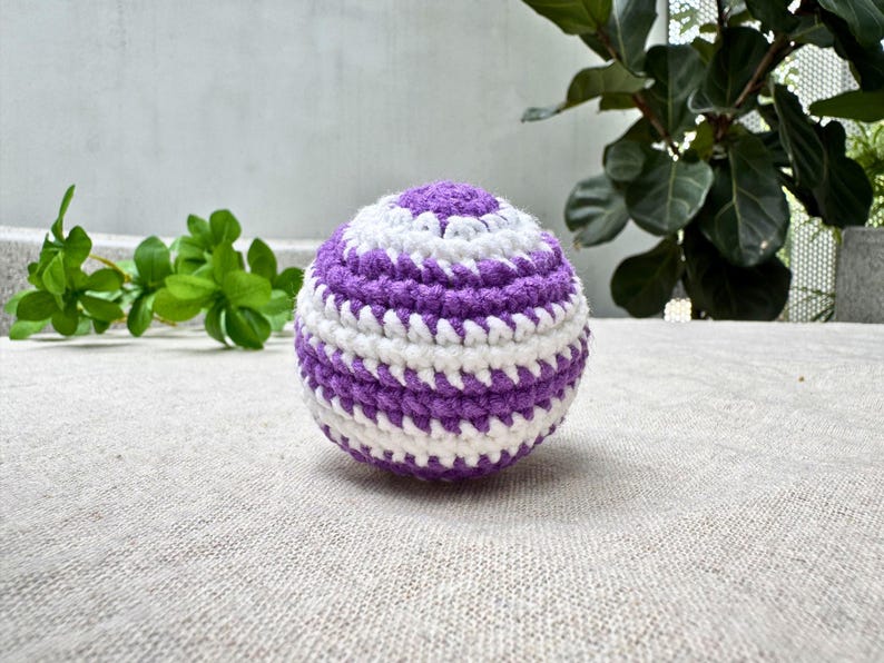 K&ouml;nnte beinhalten: Handgeh&auml;kelter Ball in Kugelform mit abwechselnden wei&szlig;en und lila Streifen. Der Ball liegt auf einer neutralfarbenen Oberfl&auml;che, im Hintergrund gr&uuml;nes Laub. Der Durchmesser betr&auml;gt ca. 7 cm.
