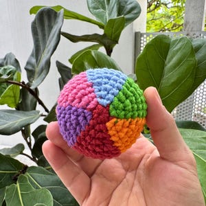 K&ouml;nnte beinhalten: Ein mehrfarbiger, geh&auml;kelter Ball mit Segmenten in Rosa, Blau, Gr&uuml;n, Orange, Lila und Rot. Der Ball wird in einer Hand gehalten, mit einem Hintergrund aus gr&uuml;nen Bl&auml;ttern und einer wei&szlig;en Wand.