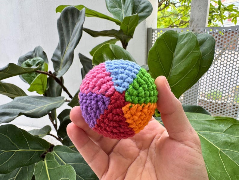 K&ouml;nnte beinhalten: Ein mehrfarbiger, geh&auml;kelter Ball mit Segmenten in Rosa, Blau, Gr&uuml;n, Orange, Lila und Rot. Der Ball wird in einer Hand gehalten, mit einem Hintergrund aus gr&uuml;nen Bl&auml;ttern und einer wei&szlig;en Wand.