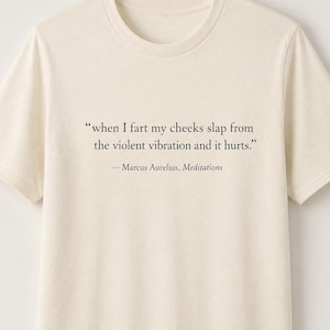 May include: A cream-colored short-sleeve t-shirt featuring black printed text that reads: "when I fart my cheeks slap from the violent vibration and it hurts." followed by the attribution "— Marcus Aurelius, Meditations" in a serif font.