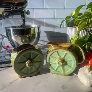 May include: Two ceramic vessels with a light green glaze and a dark brown rim, each featuring a molded wagon wheel design on the side. One vessel is a pitcher with a handle and a spout, while the other is a bowl-shaped container with an open top.