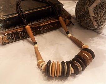 May include: A wooden necklace with brown and beige beads. The beads are stacked in a circular pattern and are interspersed with white beads. The necklace is lying on a table with a book and a vase in the background.