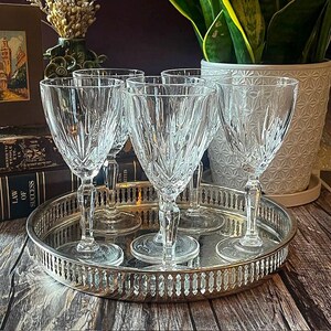 May include: A set of five clear crystal wine glasses with a textured design, displayed on a silver-colored, round tray. The tray has a decorative edge. The glasses are arranged on a wooden surface.