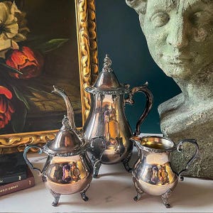 May include: A silver tea set with ornate detailing, including a teapot, creamer, and sugar bowl. The set is displayed on a white surface, with a painting and a statue in the background. The tea set reflects the surrounding objects.