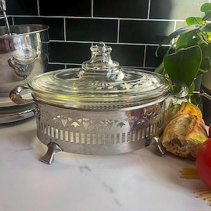 May include: A vintage glass casserole dish with a clear glass lid and a silver metal stand. The stand has decorative cutouts and small feet. The lid has a floral design. The dish is on a white surface.