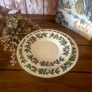May include: Two white ceramic plates, one stacked on the other, featuring a green and red holly leaf design around the rims. The plates are set on a dark wooden surface, with a Christmas card visible in the background.