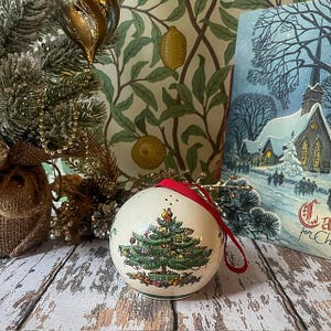 May include: A round, cream-colored ceramic Christmas ornament featuring a detailed illustration of a decorated pine tree with gifts underneath and a figure at the top. The ornament has a small red ribbon loop at the top for hanging.
