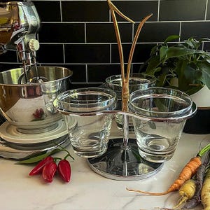 May include: A chrome and gold-toned serving piece with four clear glass cups. The stand has a decorative gold handle. Red peppers and carrots are in the foreground. A stainless steel mixer is in the background.