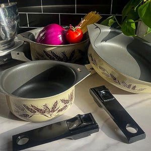 May include: A set of four vintage cookware pots with a wheat design. The pots are cream colored with brown wheat stalks and have black handles. The pots are sitting on a light brown surface with a red onion and a red tomato in one of the pots.