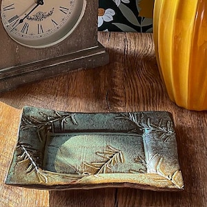 May include: Rectangular ceramic dish with a raised botanical design in shades of green and brown. The dish has a textured surface and is likely used for serving or display. The design features a detailed leaf pattern.