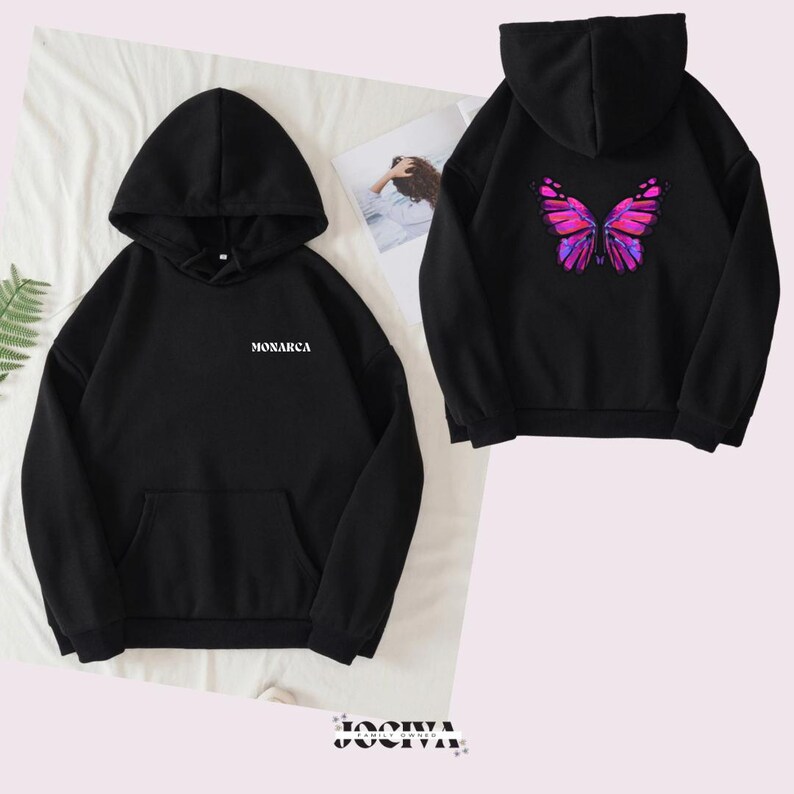 Puede incluir: Sudadera con capucha negra con bolsillo delantero y capucha. La parte delantera presenta la palabra "MONARCA" en blanco. La parte trasera de la sudadera con capucha muestra un vibrante dise&ntilde;o de mariposa rosa y morado. La sudadera con capucha se muestra sobre una superficie blanca.