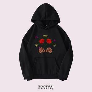 Puede incluir: Sudadera con capucha negra con bolsillo delantero. El dise&ntilde;o gr&aacute;fico presenta dos rosas rojas, pu&ntilde;os con tatuajes y el texto "SAUCE BOYZ". El dise&ntilde;o tambi&eacute;n incluye mariposas y s&iacute;mbolos circulares.