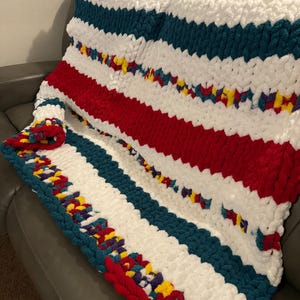 Puede incluir: Una manta de punto grueso con rayas blancas, rojas, azul verdoso y un hilo multicolor con detalles amarillos, rojos, morados y azules.