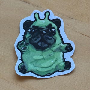 May include: A sticker featuring a green pug-like creature with black facial features and small antennae. The cartoonish design has a white border and is set against a light wood grain background.