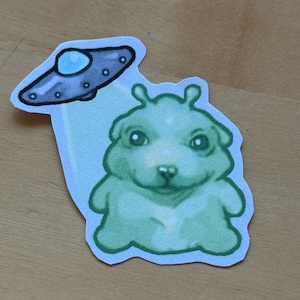 May include: A sticker featuring a green alien-dog hybrid with large eyes and a small nose. Above the alien is a gray and blue UFO. The sticker has a white border and a light blue background.