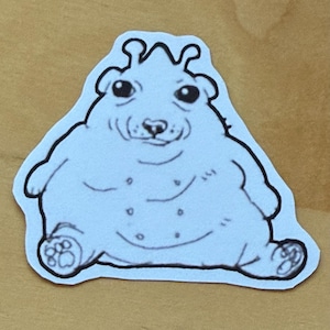 May include: A light blue sticker featuring a cartoon bear-like creature with a round body, small eyes, and a smiling expression. The creature has a black outline and is sitting down. The sticker is on a tan background.