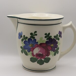 May include: A cream-colored ceramic pitcher featuring a hand-painted floral motif with a large central magenta rose, smaller blue and purple flowers, and green leaves. The pitcher has a dark green rim and base trim, a curved handle, and a spout.