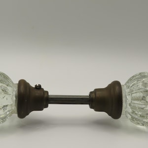 May include: A door knob set featuring two clear, fluted glass knobs attached to a central metal spindle. Each glass knob is connected to a dark, aged bronze-toned metal base, with a small visible screw on one of the circular mounting collars.