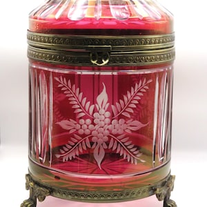 May include: A cylindrical ruby red glass casket featuring vertical cut panels and an etched floral motif on the front. The container is encased in a brass frame with a hinged lid and rests on four ornate, claw-shaped brass feet at the base.