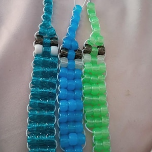 May include: Three beaded bracelets in teal, blue, and green. Each bracelet has a central design with black, white, and clear beads. The bracelets are strung on white cord.