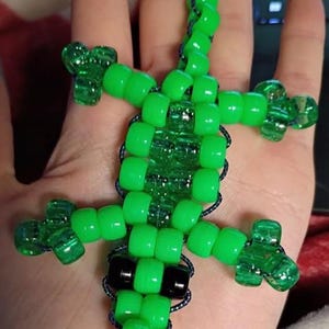 May include: A beaded lizard figurine in bright green, black, and clear beads. The lizard is made with a series of round beads strung together with dark blue string, with clear beads for the feet. The lizard is held in a hand.
