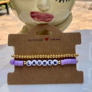 May include: A set of two bracelets with the name "LAUREN" spelled out in white beads on a purple beaded bracelet. The bracelets are on a brown card with the text "Handmade with love".