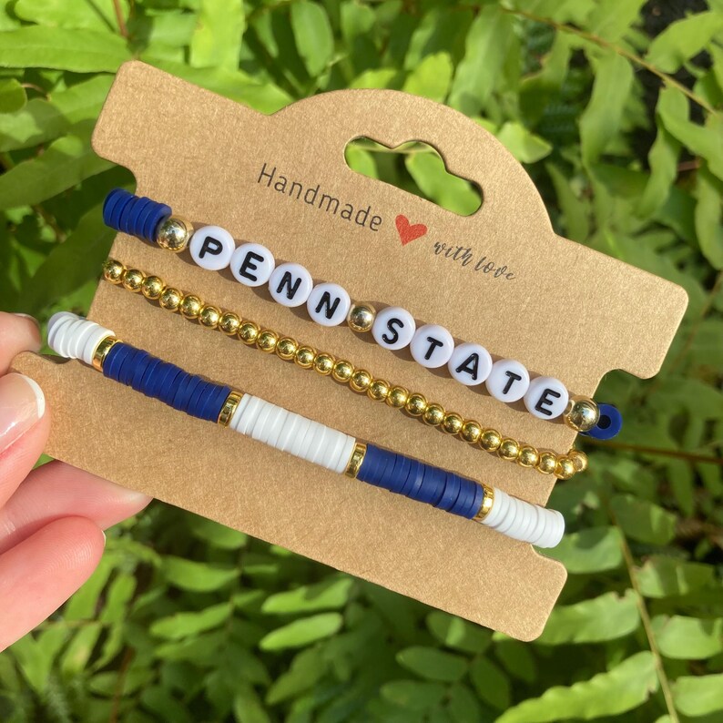 May include: Three bracelets with blue and white beads and gold beads spelling out "Penn State" with the words "Handmade with love" on a brown card.