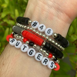 May include: A stack of bracelets with white beads that spell out "RUTGERS" and black and silver beads. There are also two white beads with the numbers "#46" on them.