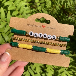 May include: Three bracelets with green and yellow beads and a white beaded word "GATORS" in the center. The bracelets are displayed on a brown card with the text "Handmade with love".