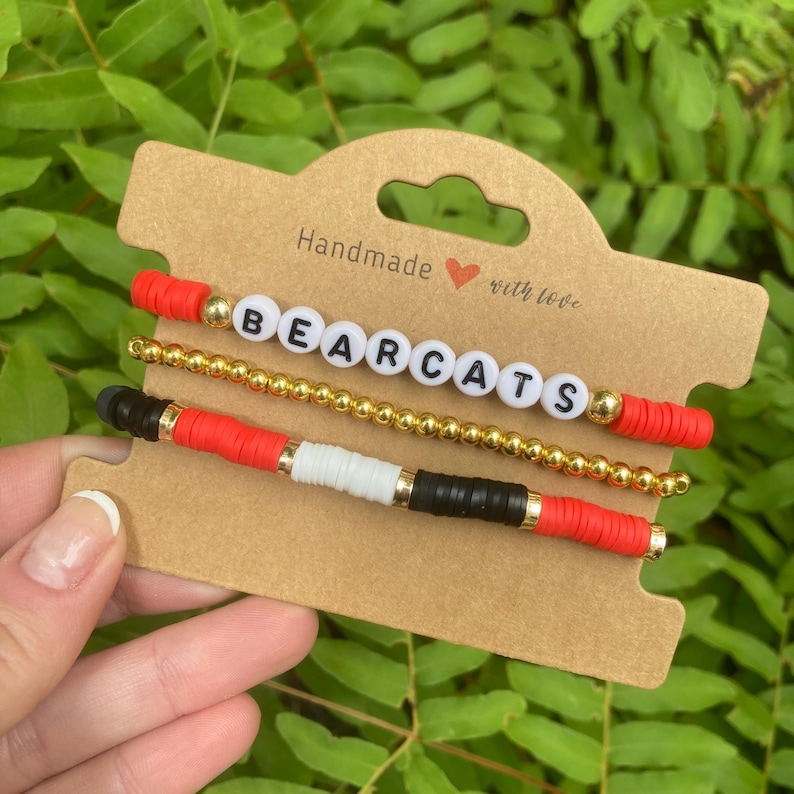 May include: Three beaded bracelets with the words "BEARCATS" in white letters on a gold chain. The bracelets are stacked on a brown card with the text "Handmade with love".