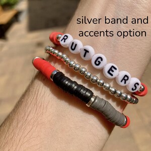 May include: A set of three bracelets, one with the word "RUTGERS" spelled out in white beads, one with silver beads, and one with red, black, and gray beads. The bracelets are on a person's wrist.