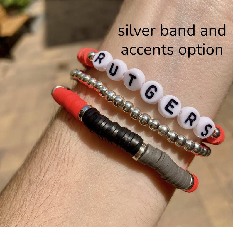 May include: A set of three bracelets, one with the word "RUTGERS" spelled out in white beads, one with silver beads, and one with red, black, and gray beads. The bracelets are on a person's wrist.