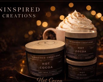 Puede incluir: Imagen de tres velas de cera de soja etiquetadas como "Hot Cocoa" con tapas de madera, apiladas sobre una bandeja dorada. Una taza de chocolate caliente con crema batida está en el fondo. El texto "Ninspired Creations" es visible.
