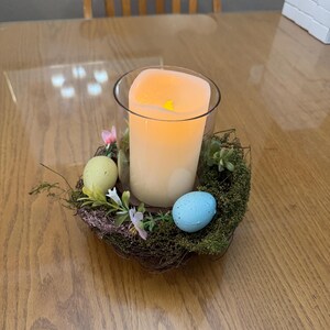 May include: A clear glass candle holder with a white candle inside, surrounded by a nest of twigs and moss. There are three decorative Easter eggs, one yellow, one light blue, and one dark blue, in the nest.