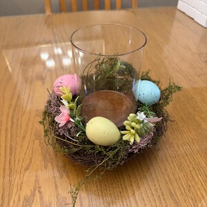 May include: A glass cylinder vase with a brown wooden base is surrounded by a nest of twigs and moss. The nest is decorated with three pastel-colored Easter eggs and small pink and white flowers.