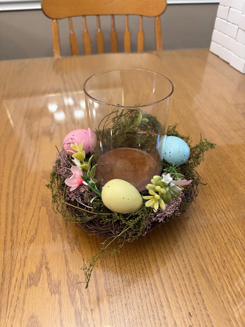 May include: A glass cylinder vase with a brown wooden base is surrounded by a nest of twigs and moss. The nest is decorated with three pastel-colored Easter eggs and small pink and white flowers.