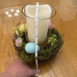 May include: A clear glass candle holder with a white candle inside, surrounded by a nest of moss, twigs, and faux eggs. The nest is decorated with pink and yellow flowers. A measuring tape is placed next to the candle holder, measuring 7 inches in height.