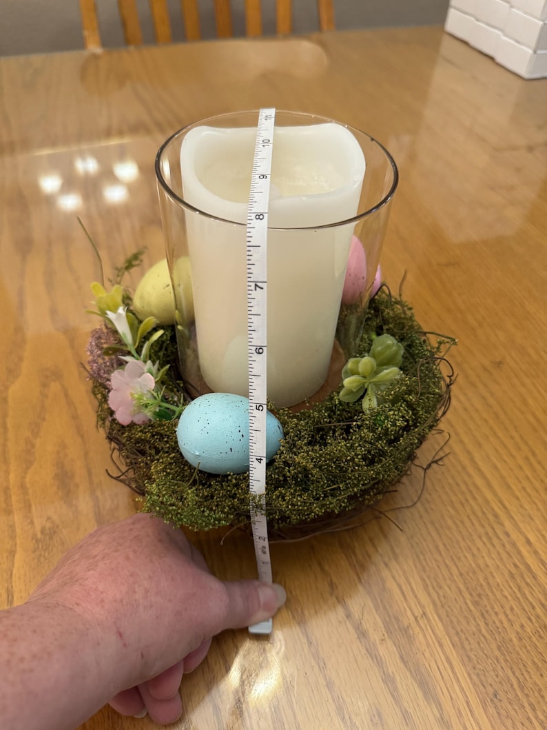 May include: A clear glass candle holder with a white candle inside, surrounded by a nest of moss, twigs, and faux eggs. The nest is decorated with pink and yellow flowers. A measuring tape is placed next to the candle holder, measuring 7 inches in height.