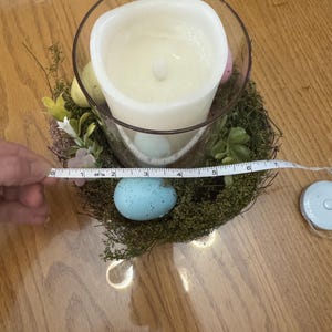 May include: A white candle in a clear glass jar surrounded by a nest of moss and faux eggs. The nest is decorated with small white flowers and green leaves. A measuring tape is placed around the nest.