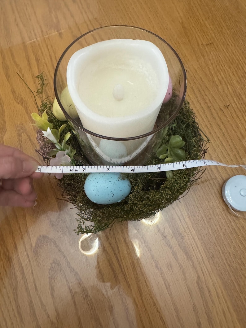 May include: A white candle in a clear glass jar surrounded by a nest of moss and faux eggs. The nest is decorated with small white flowers and green leaves. A measuring tape is placed around the nest.